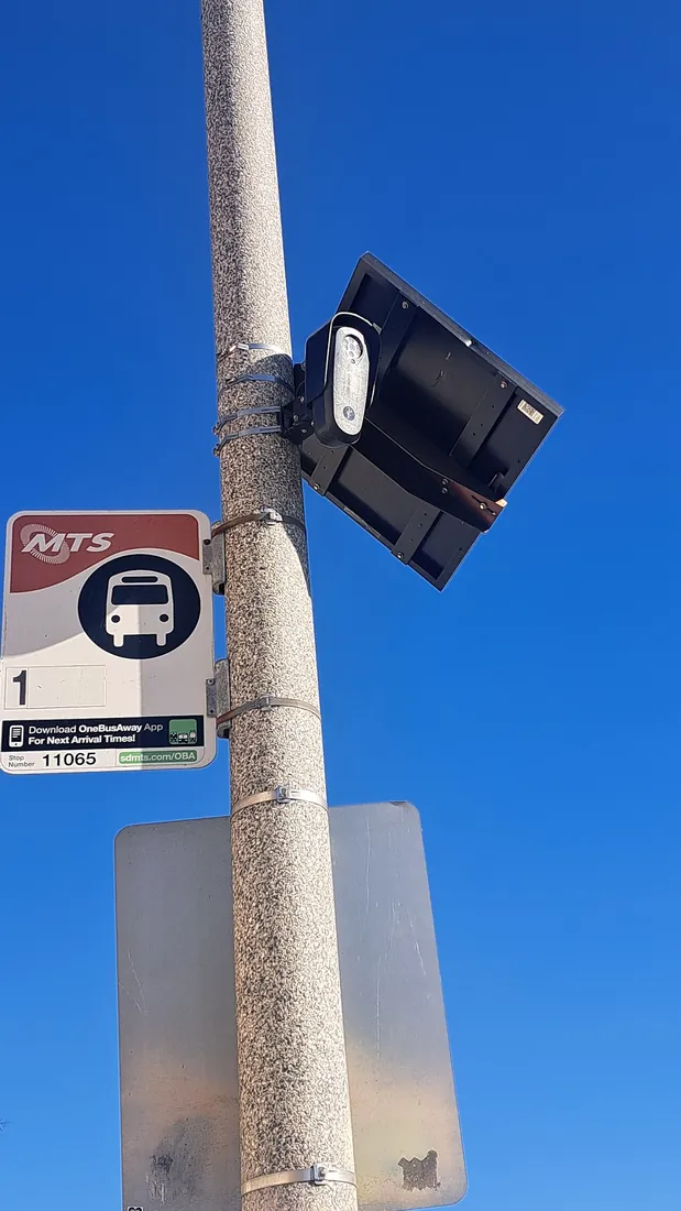 A Flock Safety license plate reader and its solar panel strapped to a pole above an MTS Route 1 bus stop sign.