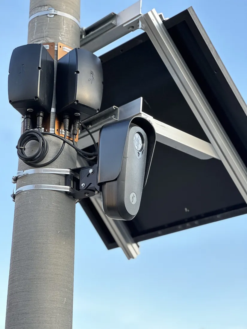 Close view of a Flock Safety camera on a gray street-light pole, below a tilted solar panel, with two black equipment boxes and cabling strapped beside it.