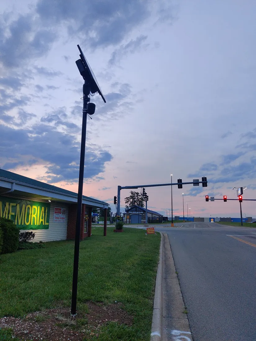 A Flock Falcon plate reader on a slim black pole with a solar panel at the top, at the edge of a lot facing a road at dusk.
