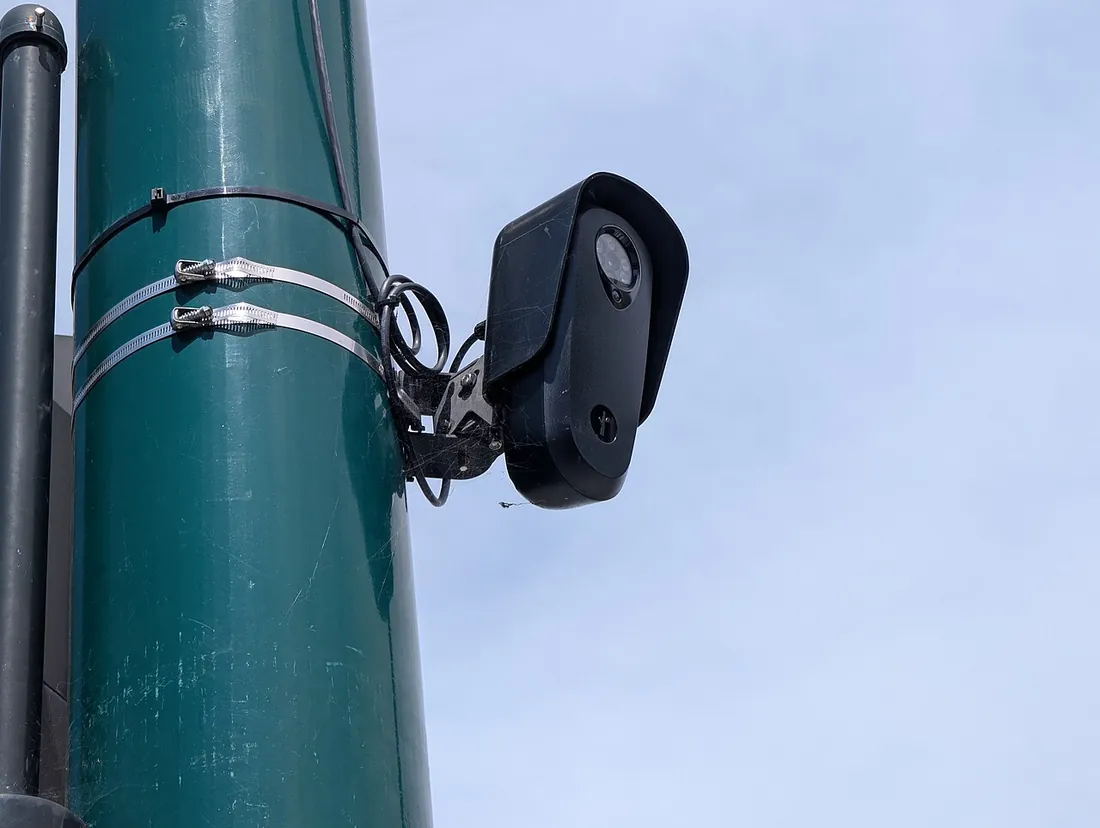 A black Flock Safety camera clamped with metal bands to a green street-light pole, with a coil of cable at the mount.