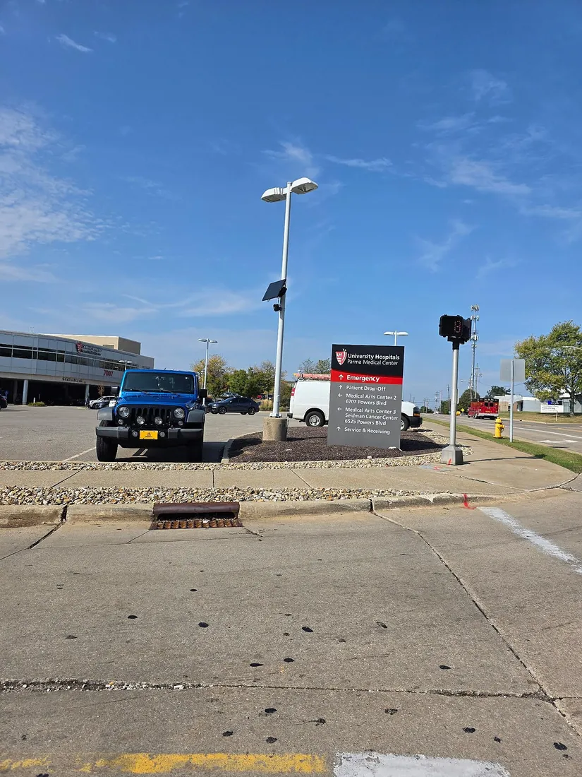 A license plate reader with a small solar panel on a light pole at the entrance to University Hospitals Parma Medical Center, next to the hospital's direction sign.
