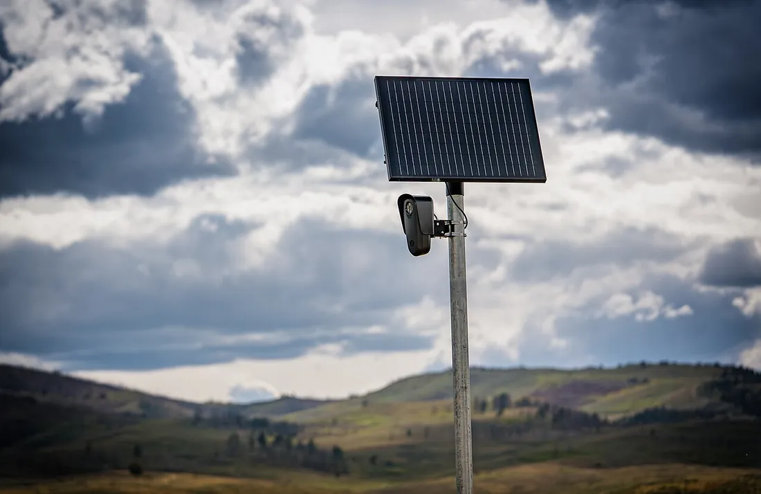 A Flock Safety camera under a solar panel on a metal post beside a rural highway, with green hills and storm clouds behind it.