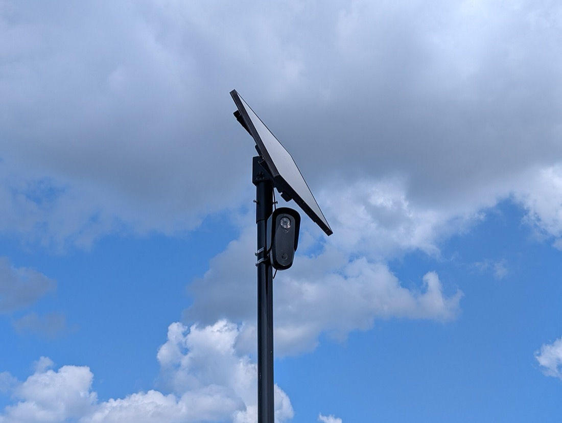 A Flock Safety license plate reader mounted on a pole with a solar panel above it, in a retail parking lot in Eatontown, New Jersey.