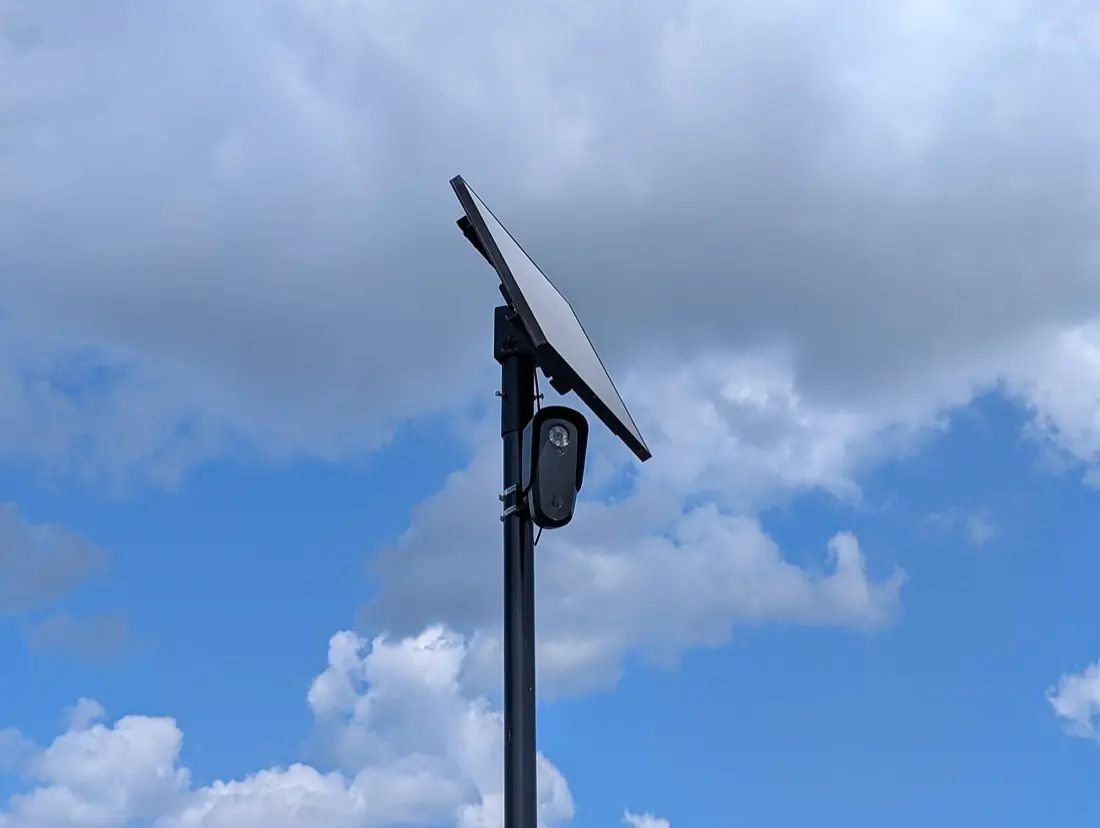 A Flock Safety license plate reader mounted on a pole with a solar panel above it, in a retail parking lot in Eatontown, New Jersey.