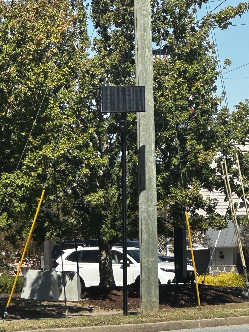 A Flock Safety license plate reader on a slim black pole beside a residential street, with a small solar panel mounted above the camera housing and no cable running to the ground.